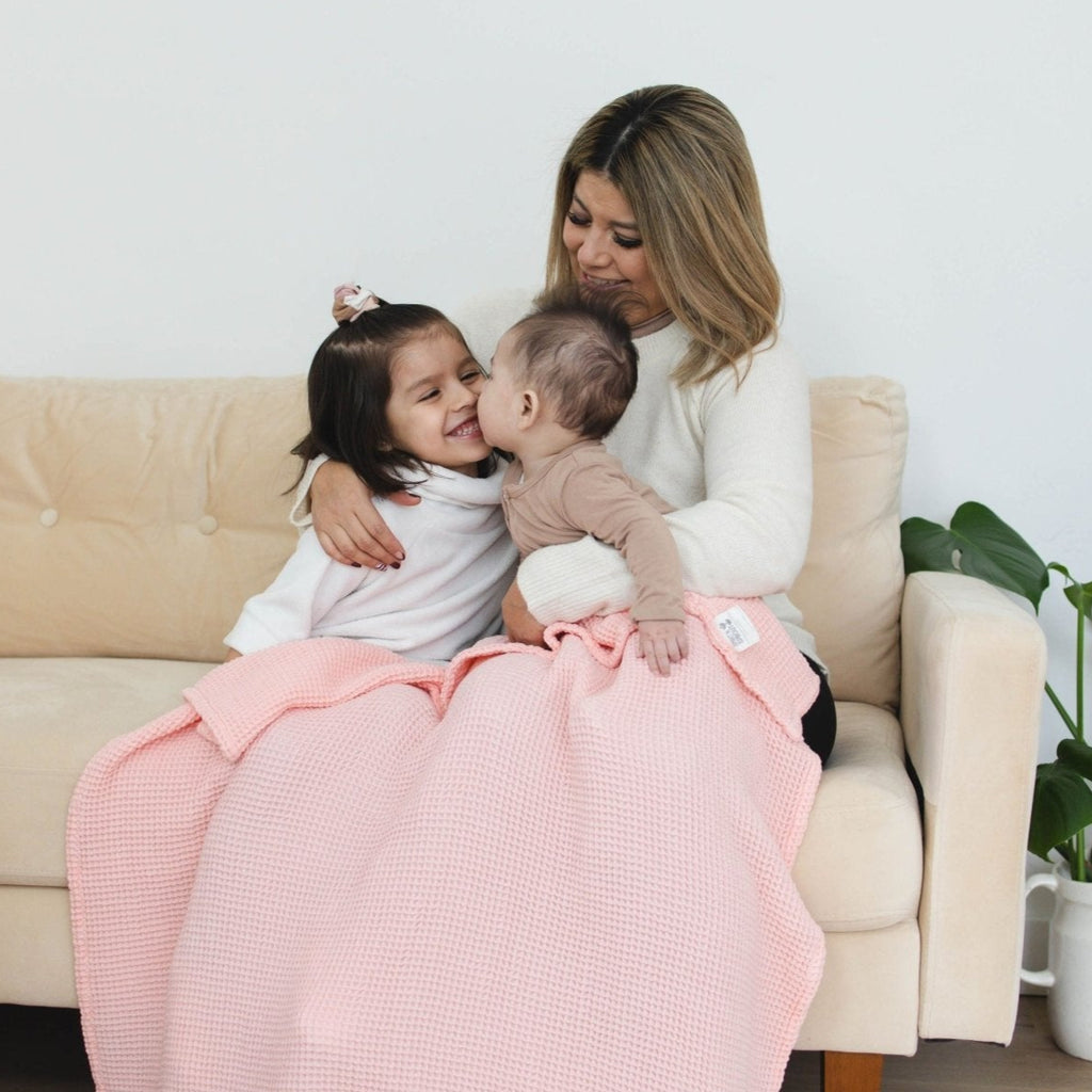 Mom on couch watches as older sibling receives a kiss from her baby sibling in a colorful blanket.